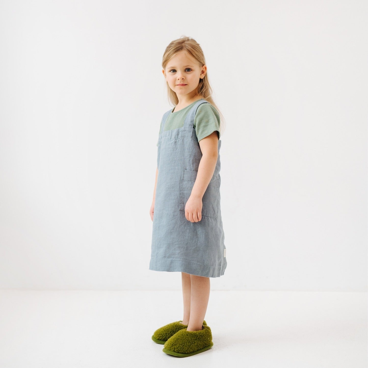 blue fog linen pinafore apron on a child in a sunlit kitchen, hands in pockets