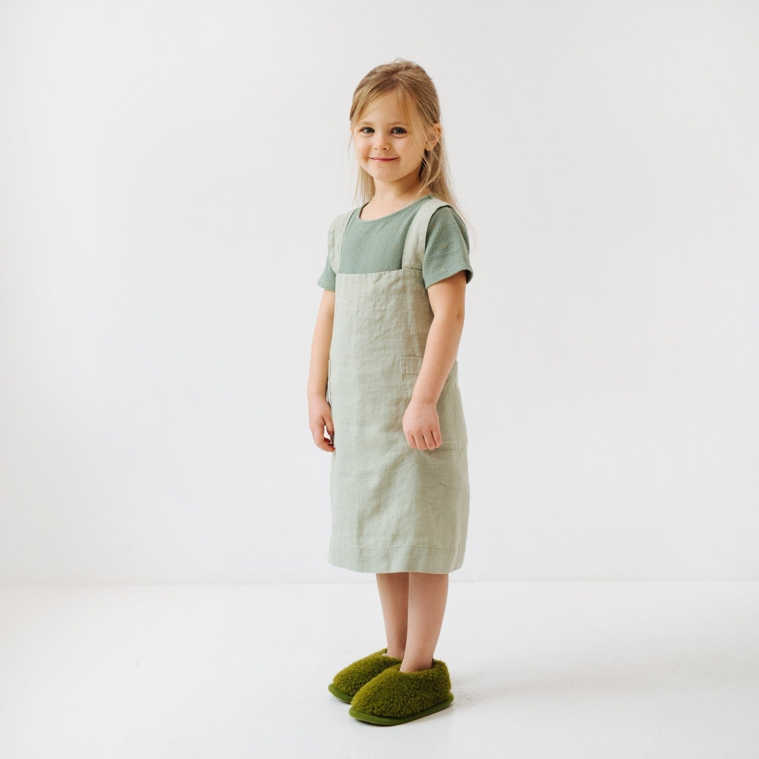 a child wearing a soft sage linen pinafore apron, smiling warmly in soft morning light