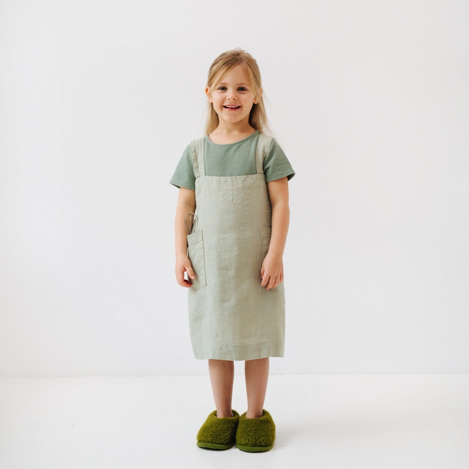 a child laughing while wearing a sage linen pinafore apron, hands in the pockets ready for a baking adventure
