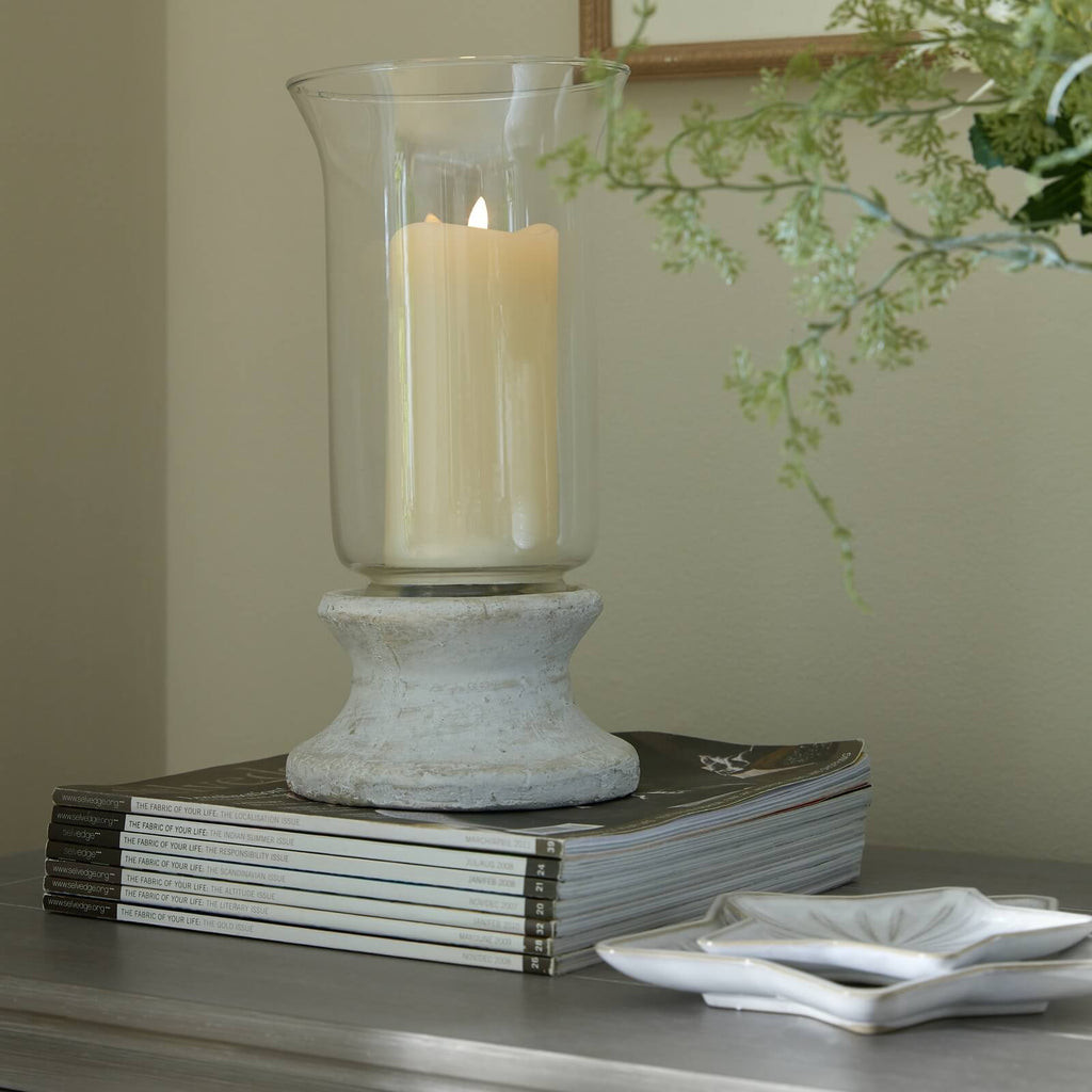 Handcrafted cream hurricane lantern with stone-effect base and antiqued patina, casting warm LED candlelight glow. by Sostter at brixbailey.com