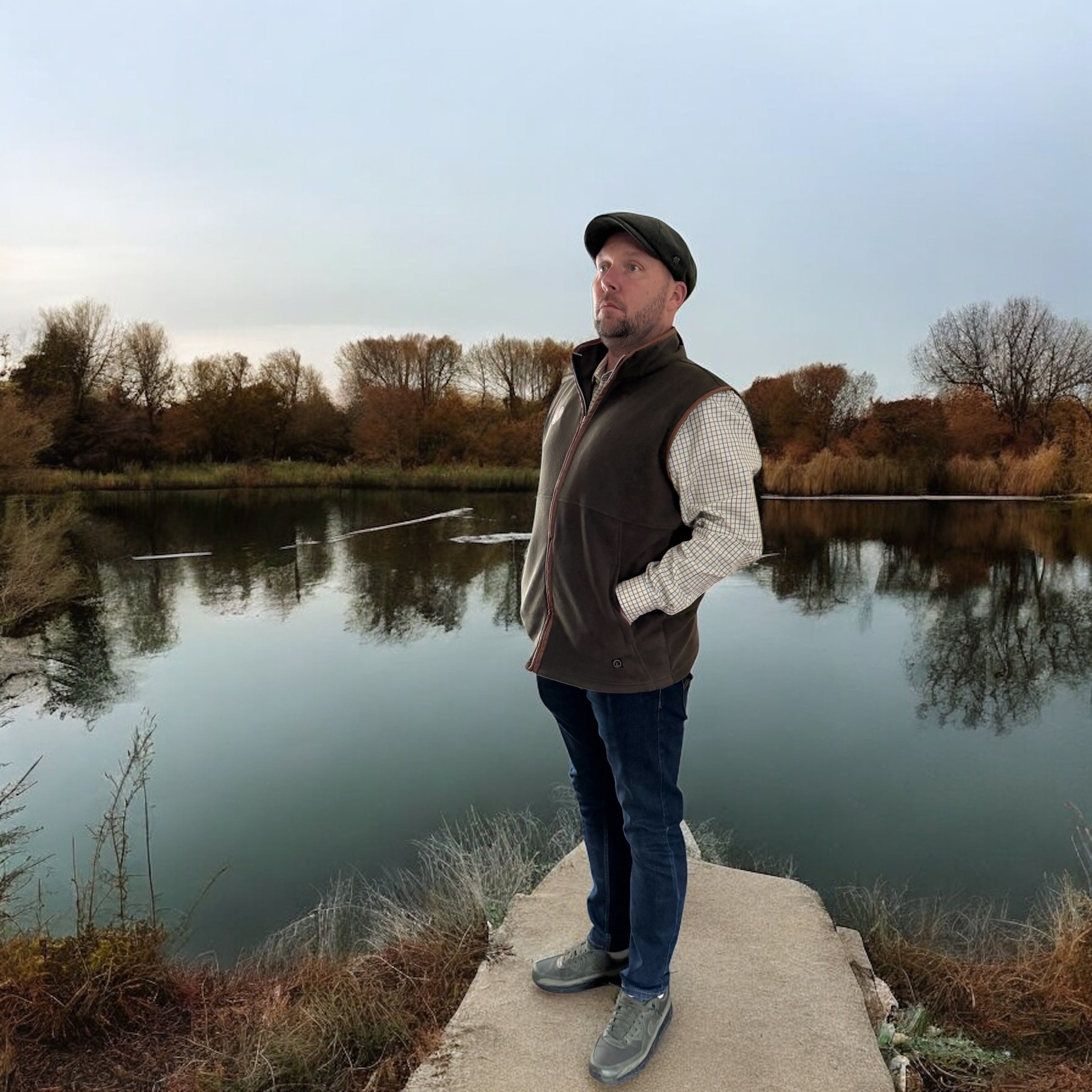 Man standing on a concrete ledge by a lake with trees in the background wear aqua lilium clothing brand on Sostter