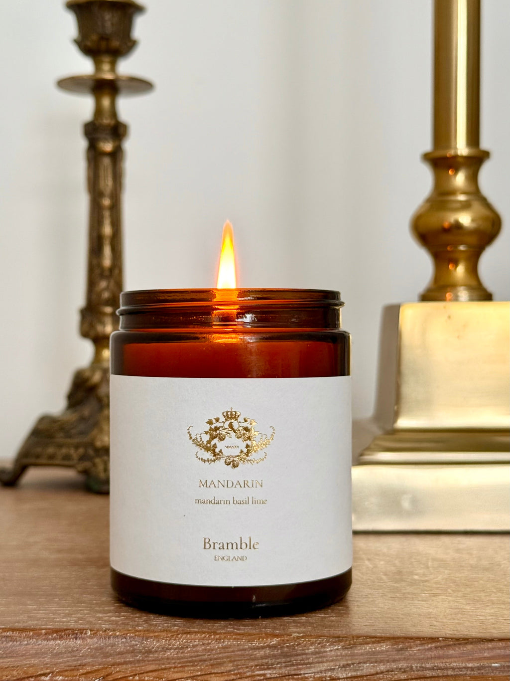 Candle in a jar with a white label featuring a logo and text, on a wooden surface with gold candle holders.