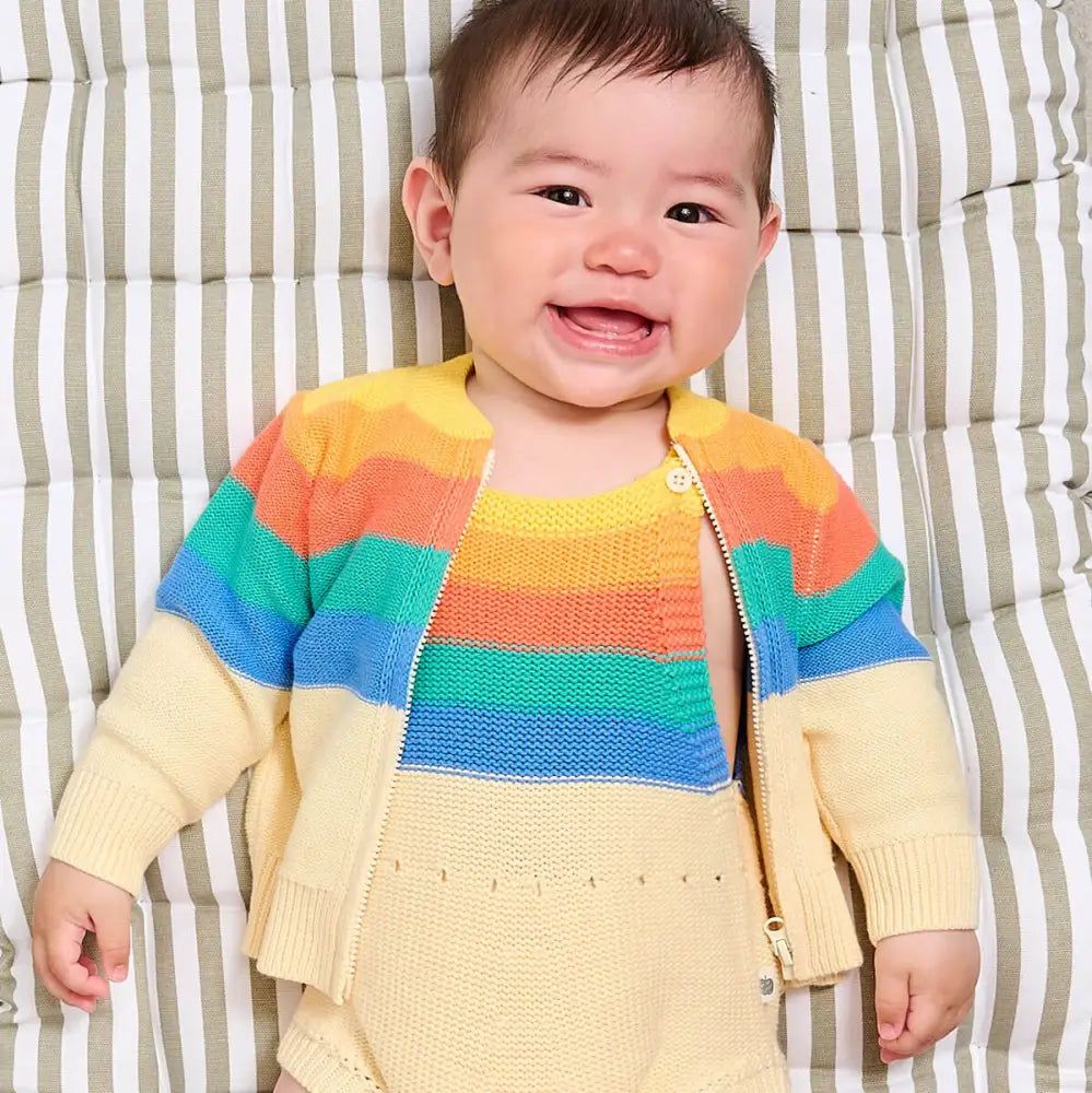 Baby wearing a colorful striped sweater over a yellow onesie on a striped background
