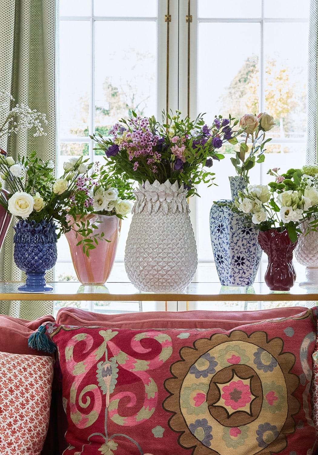 Decorative vases with flowers on a windowsill next to a colorful pillow.