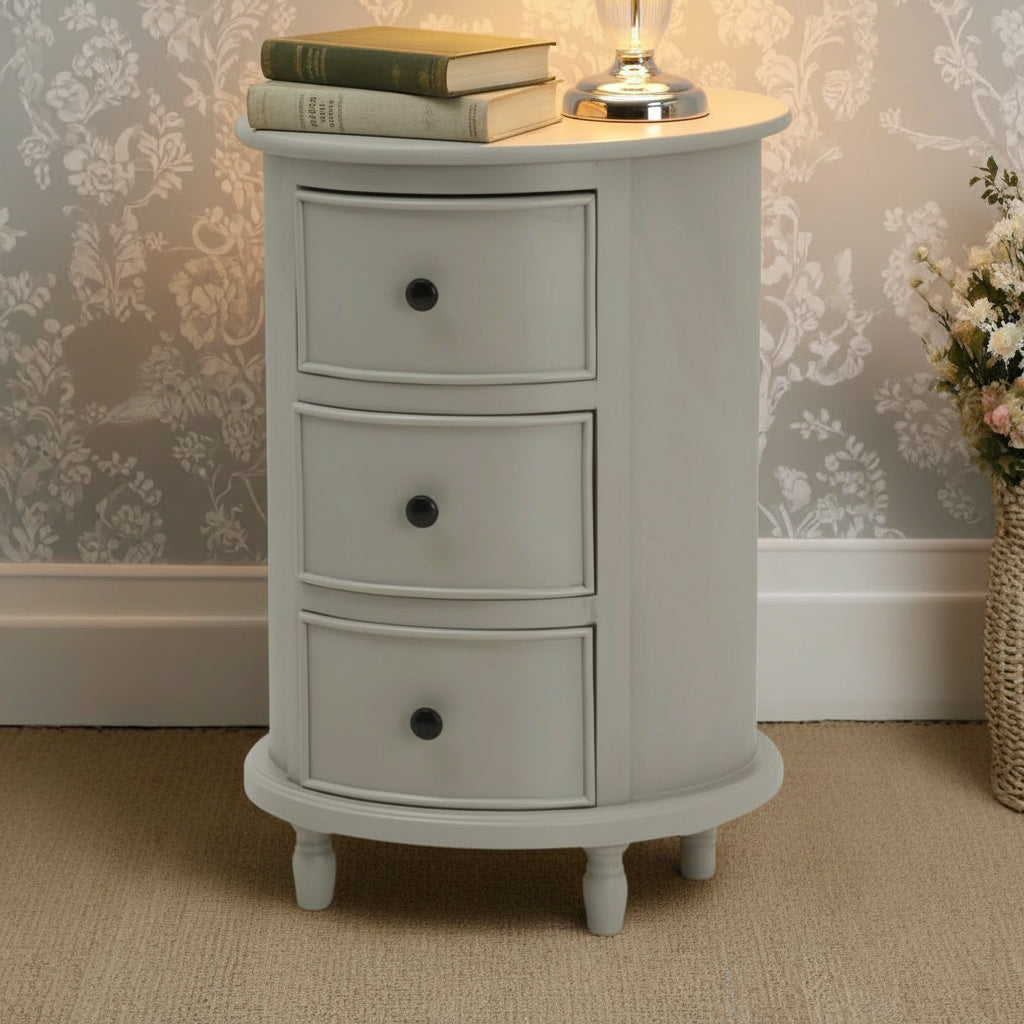 Round white nightstand with three drawers against a floral wallpapered wall.