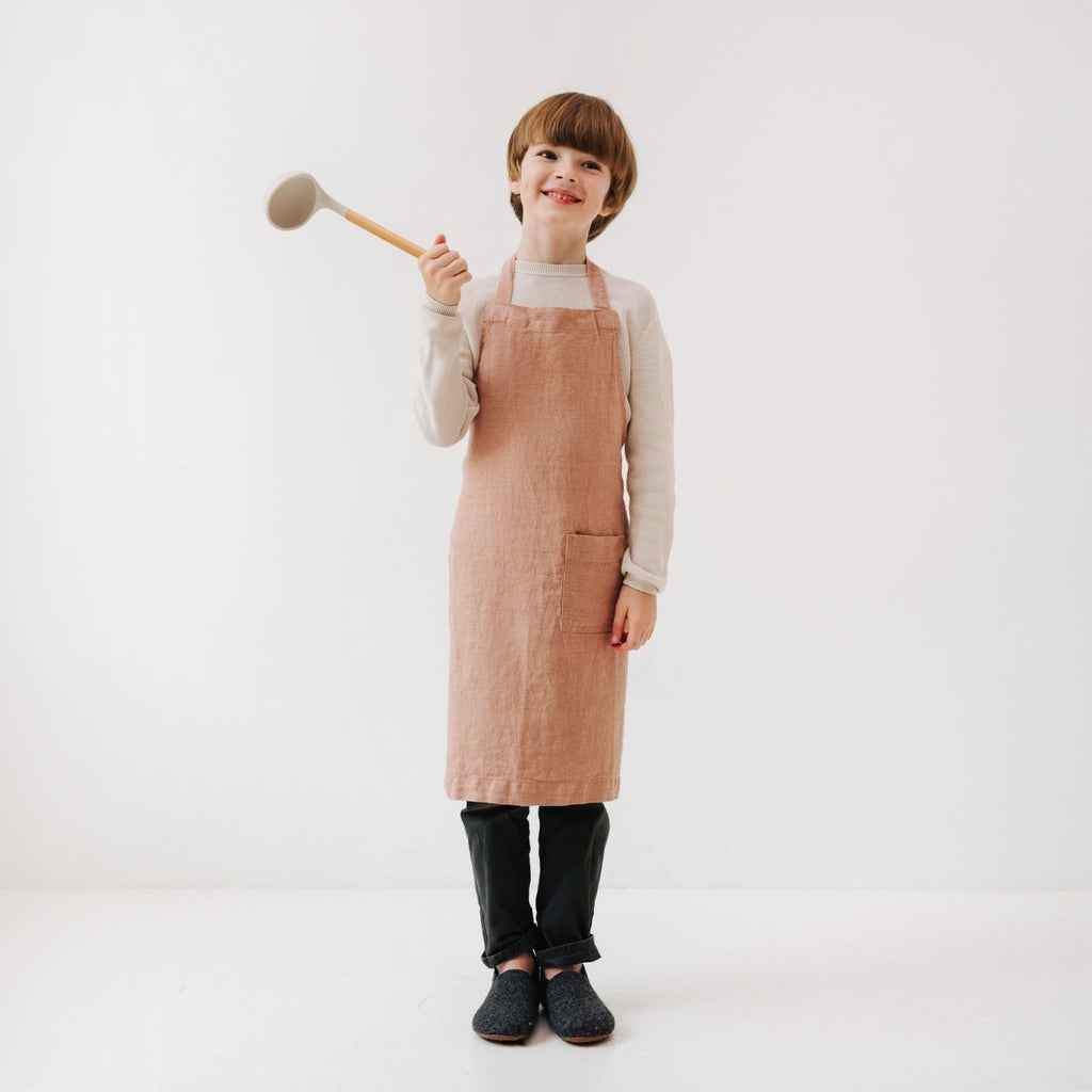 playful cafe creme linen apron for kids with front pocket, tied around a smiling child in a sunlit kitchen