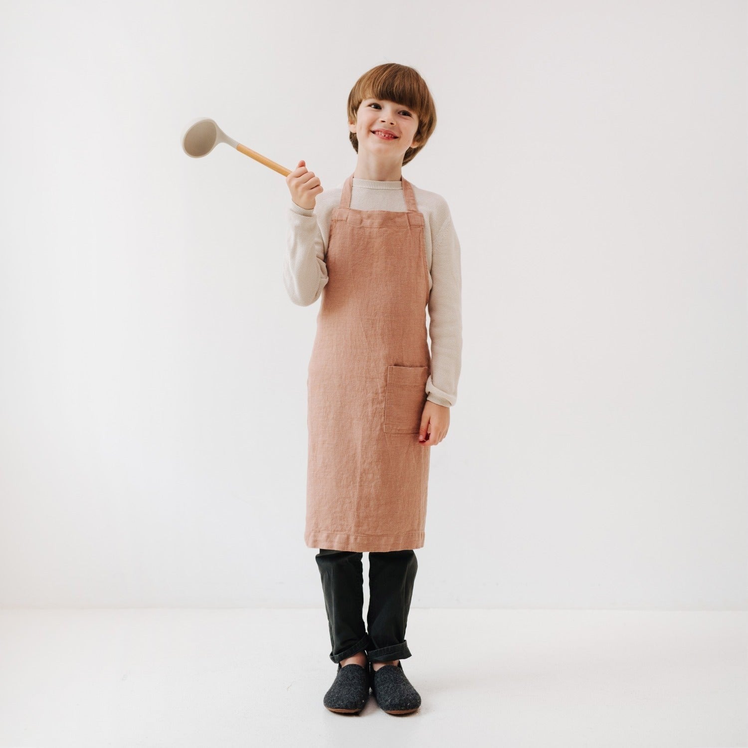 playful cafe creme linen apron for kids with front pocket, tied around a smiling child in a sunlit kitchen