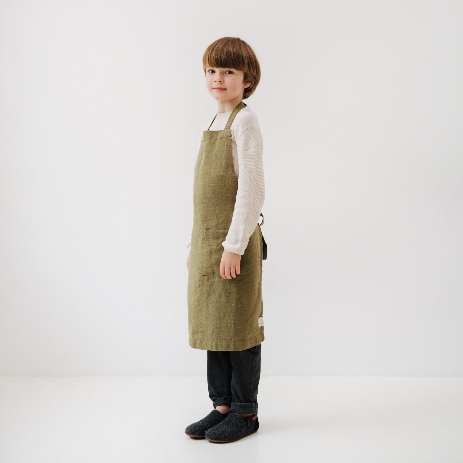 soft olive linen kids apron with pocket, gently draped on a wooden chair in warm sunlight