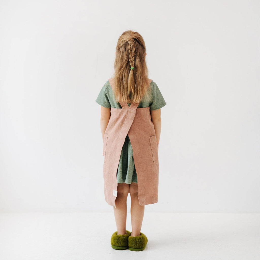 soft honey-colored linen kids apron with pockets, draped gently on a wooden chair in warm sunlight