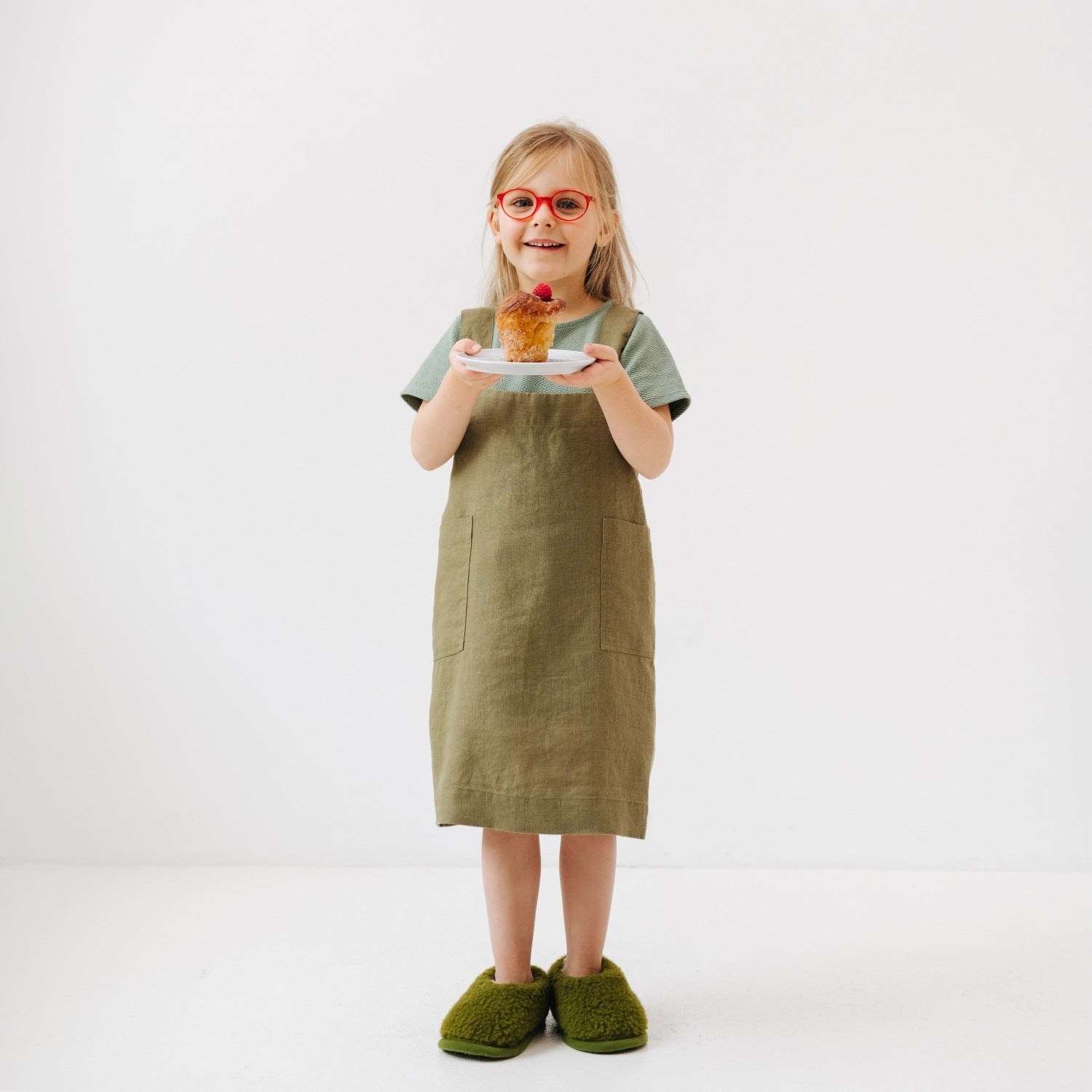 elegant olive linen pinafore apron for kids with front pockets, draped on a wooden chair in soft morning light