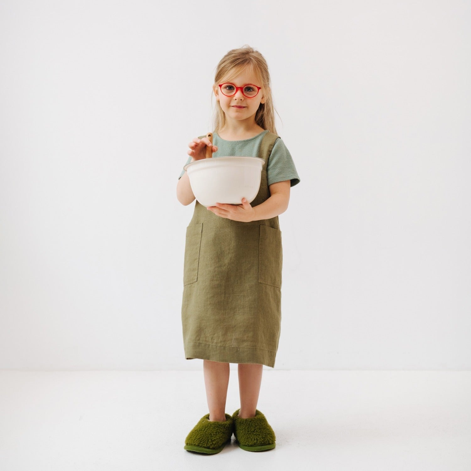 soft olive linen kids apron with pockets, draped gently on a wooden chair in warm sunlight