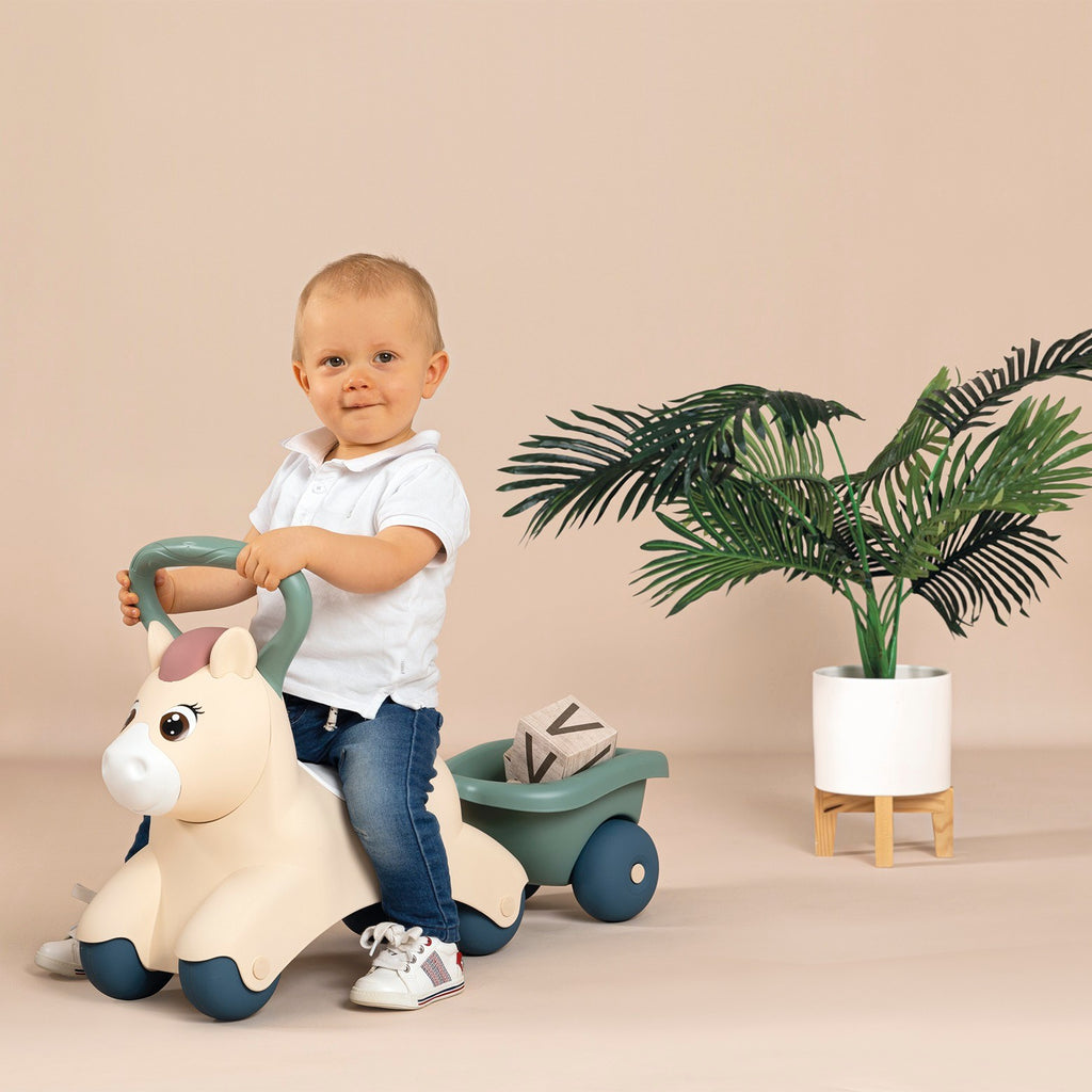 adorable toddler riding pastel pony with attached trailer for toys in sunny backyard