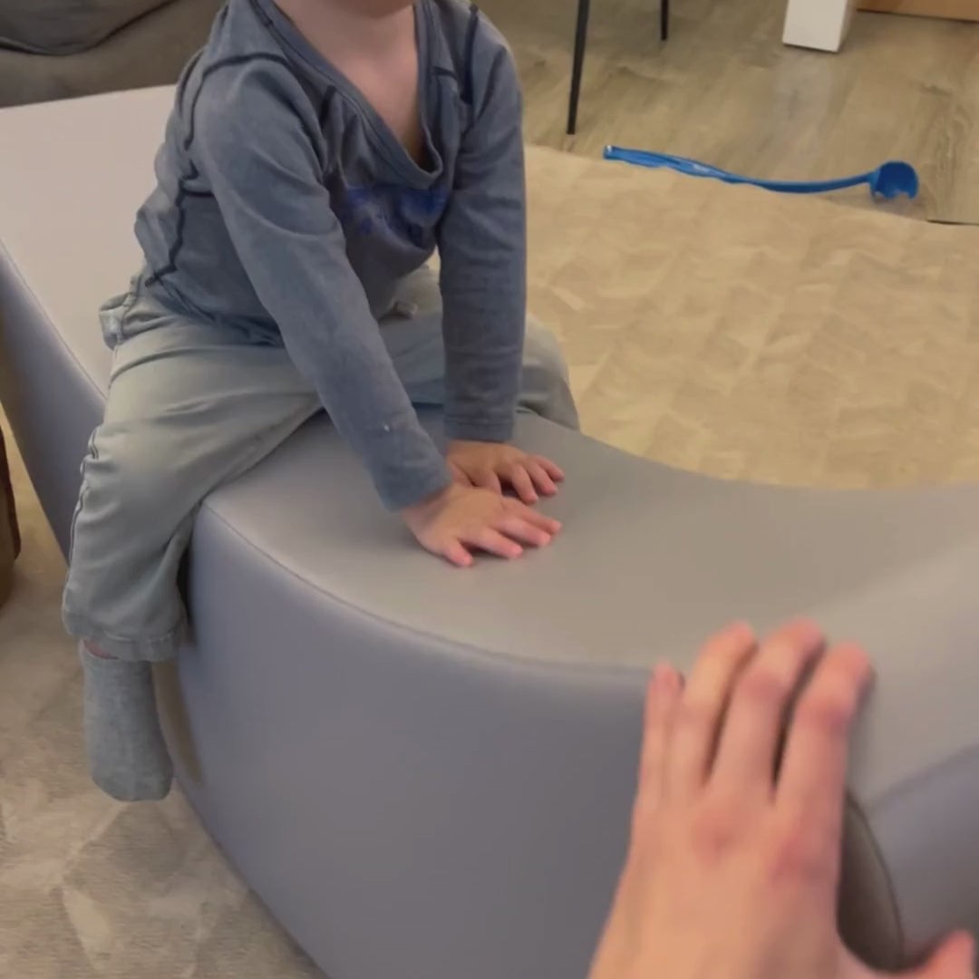 smiling toddler rocking on grey banana toy, giggling while balancing on soft eco-leather foam