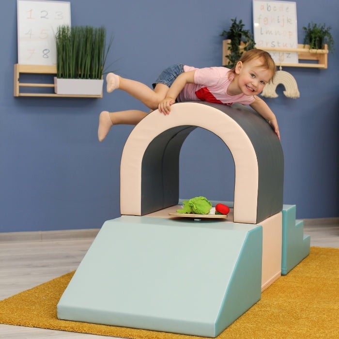 smiling toddler crawling through pastel soft play tunnel, giggling with friends in a cozy playroom
