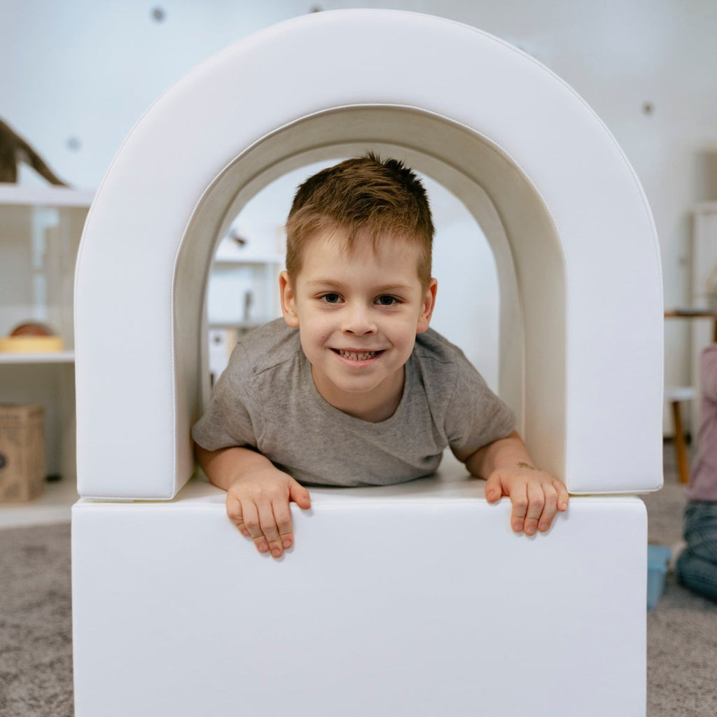 smiling toddler crawling through a bright white soft play tunnel, giggling with friends