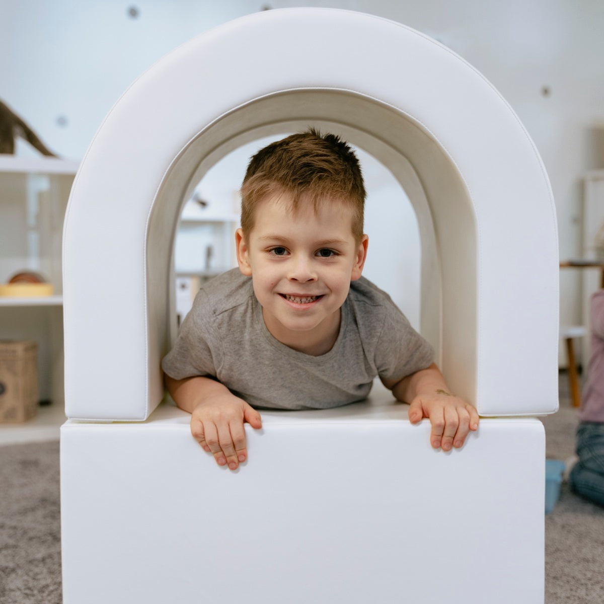 smiling toddler crawling through a bright white soft play tunnel, giggling with friends