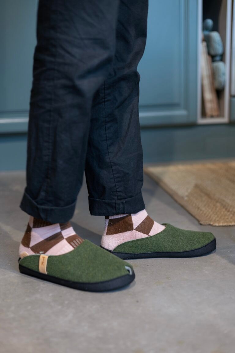 soft moss green lambswool slippers with black microfibre sole, resting on a cozy wooden floor