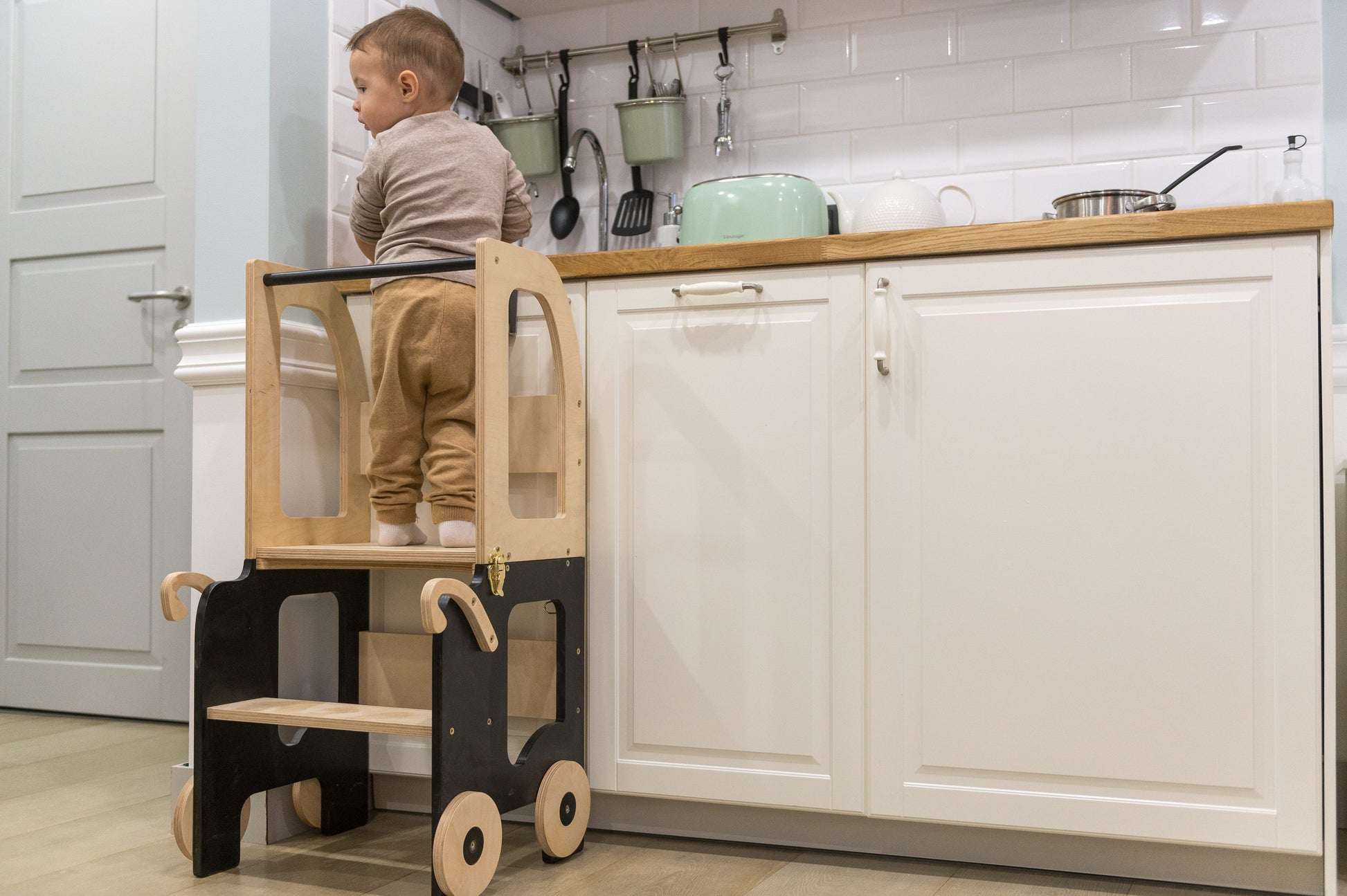 a gleeful toddler in a golden yellow kitchen helper tower, reaching for a mixing bowl with floury hands